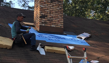 Roof repair near chimney with worker installing weatherproofing sheet.