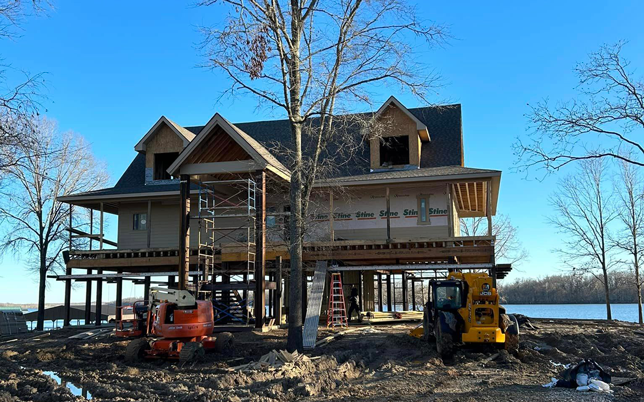 Elevated house under construction with scaffolding and machinery by a lakeside with clear blue sky.