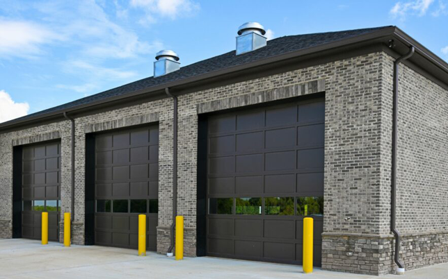 Modern brick garage building with three large black doors and yellow bollards, under a blue sky.