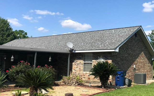 Brick house with grey shingle roof, satellite dish, and manicured garden under a blue sky with a few clouds.