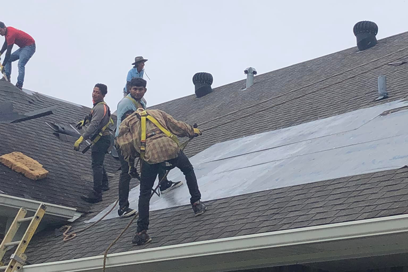 Roofers working on shingle roof installation with safety gear on a cloudy day.