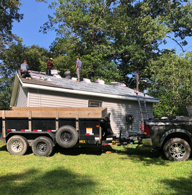 Roofers working on house with nearby truck and trailer amidst trees, sunny day, exterior home improvement project.